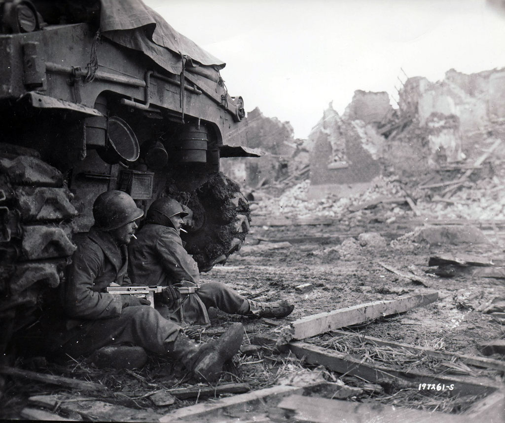 Two GIs from the 9th Infantry Division shelter beneath a Sherman tank on December 11 in the smashed German town of Geich, near Dueren.